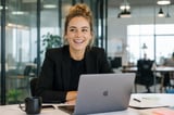 Professional woman with curly blonde hair smiling at her desk while working on a laptop